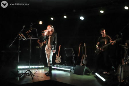 Kolorowe zdjęcie. Na scenie kobieta i dwóch mężczyzn. Kobieta śpiewa do mikrofonu. Zerka na publiczność. Za nią mężczyźni grają na gitarach.