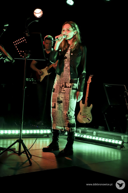 Kolorowe zdjęcie. Na scenie kobieta i mężczyzna. Kobieta śpiewa do mikrofonu, który trzyma w ręku. Zerka na publiczność. Za nią mężczyzna gra na gitarze.