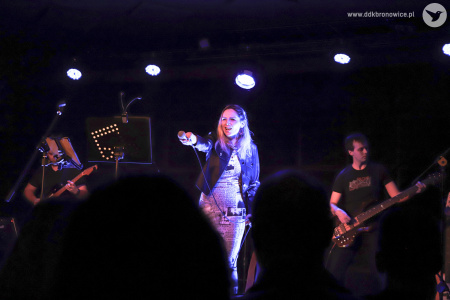 Kolorowe zdjęcie. Na scenie wokalistka zespołu. Wyciąga mikrofon w stronę publiczności. Za nią gitarzysta i basista.