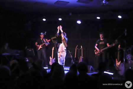 Kolorowe zdjęcie. Na scenie wokalistka zespołu klaszcze nad swoja głową. Za nią gitarzysta i basista.
