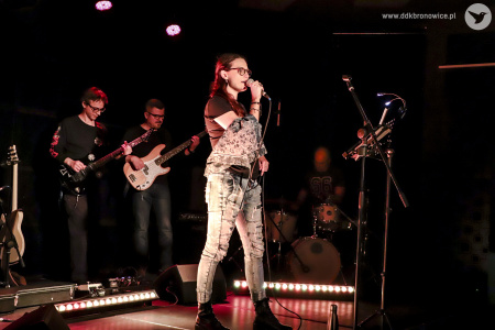 Kolorowe zdjęcie. Na scenie kobieta i trzech mężczyzn. Kobieta śpiewa do mikrofonu. Zerka na publiczność. Za nią dwóch mężczyzn gra na gitarach i jeden na perkusji.