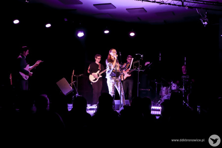 Kolorowe zdjęcie. Na scenie kobieta i czterech mężczyzn. Kobieta śpiewa do mikrofonu. Zerka na publiczność. Za nią dwóch mężczyzn gra na gitarach, jeden na basie i jeden na perkusji.