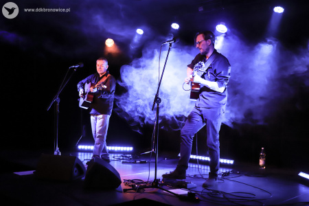 Kolorowe zdjęcie. Na zadymionej scenie muzycy: Adam Jarząbek i Jurek Jurkiewicz. Mężczyźni grają na gitarach.
