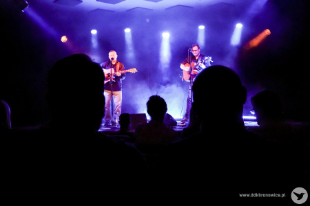 Kolorowe zdjęcie. Kadr na scenę z oddali. Muzycy grają na gitarach. Zdjęcie zza publiczności.