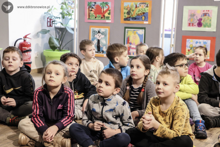 Grupa dzieci siedzi na podłodze. Dzieci widoczne na pierwszym planie siedzą po turecku.