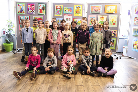 Grupa dzieci ustawiona do zdjęcia. Dzieci w pierwszym rzędzie siedzą na podłodze. Dzieci w drugim i trzecim rzędzie stoją. W tle prace plastyczne.