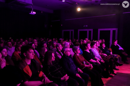 Zdjęcie przedstawia kilkadziesiąt osób siedzących na krzesłach od strony publiczności. Skierowani do widza prawym bokiem. Publiczność siedzi w poświacie różowego światła.