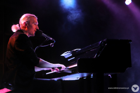 Mężczyzna ubrany na czarno, z białymi włosami związanymi w niski kucyk gra na fortepianie i śpiewa do mikrofonu.