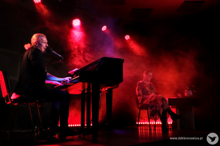 Na pierwszym planie po lewej stronie mężczyzna gra na fortepianie. Do widza skierowany jest prawym bokiem. Jest ubrany w ciemne kolory, ma białe włosy związane w kucyk. W głębi sceny, na drugim planie po prawej stronie siedzi kobieta przy małym stoliku. Scena oświetlona jest czerwonym światłem z reflektorów.