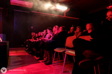 Zdjęcie dwóch rzędów siedzącej publiczności zrobione z prawej strony. Publika oświetlona czerwonym światłem. W pierwszym rządzie stoją dwa puste krzesła.