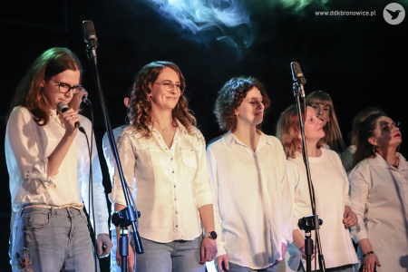 Grupa pięciu kobiet stoi w rzędzie, który przebiega przez całą szerokość zdjęcia. Kobieta najbliżej lewej krawędzi zdjęcia trzyma w dłoni mikrofon. Wszystkie ubrane są w białe bluzki i niebieskie dżinsy. Nad nimi unosi się obłok niebieskiej mgły.