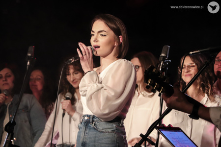 W centralnej części zdjęcia dziewczyna o brązowych włosach do ramion, w białej bluzce i niebieskich dżinsach śpiewa do mikrofonu. Za nią stoi grupa kilku chórzystek w białych bluzkach, jedna ma dodatkowo dżinsową koszulę.