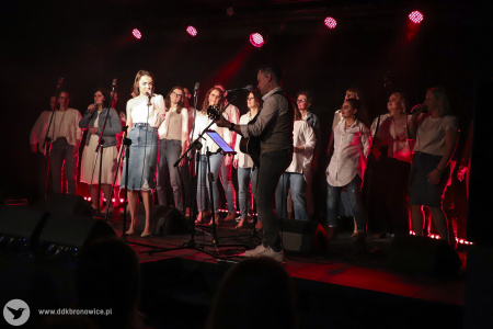 Na zdjęciu widać grupę kilkudziesięciu osób stojących na scenie. Osoby stoją w rzędzie widocznym od prawej  do lewej strony. Z przodu, bliżej lewej strony, dziewczyna w dżinsowej spódnicy do kolan śpiewa do mikrofonu. Z prawej strony mężczyzna skierowany w lewą stronę gra na gitarze. Grupę od góry oświetlają reflektory o czerwonym świetle.