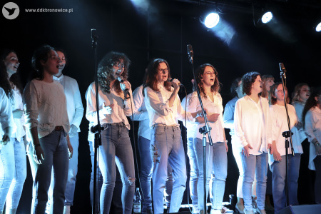 Rząd kilkunastu chórzystów. Dwie kobiety śpiewają do mikrofonów. Wszyscy ubrani w białe koszule i błękitne dżinsy.