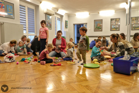 Kolorowe zdjęcie. Dzieci i rodzice siedzą na podłodze i wykonują kukiełki z drewnianej łyżki, materiałów i włóczki.