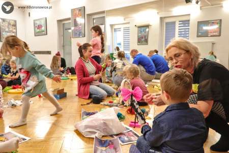 Kolorowe zdjęcie. Dzieci z rodzicami siedzą na podłodze i wykonują kukiełki. Po prawej stronie instuktorka DDK Bronowice tłumaczy coś chłopcu.