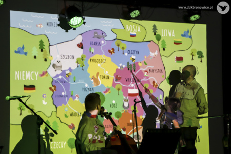 Kolorowe zdjęcie. Z tyłu sceny wyświetlona kolorowa mapa Polski. Przed mapą mężczyzna i dziecko wskazują na Kurpie Zielone.