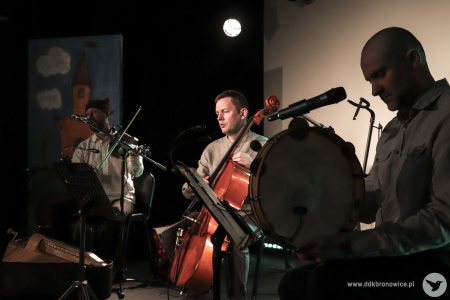Kolorowe zdjęcie. Na scenie kameralnej w Pracowni Scenicznej w Pracowniach Kultury Maki siedzi trzech mężczyzn z instrumentami: skrzypce, bęben obręczowy, wiolonczela.