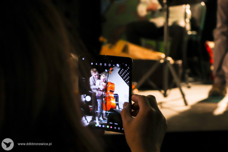 Kolorowe zdjęcie. Kadr na kobietę robiącą zdjęcie telefonem. Na ekranie telefonu widać dziewczynkę i mężczyznę na scenie. Mężczyzna uczy dziewczynkę grać na wiolonczeli.