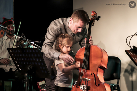Kolorowe zdjęcie. Dziewczynka i mężczyzna na scenie. Mężczyzna uczy dziewczynkę grać na wiolonczeli.