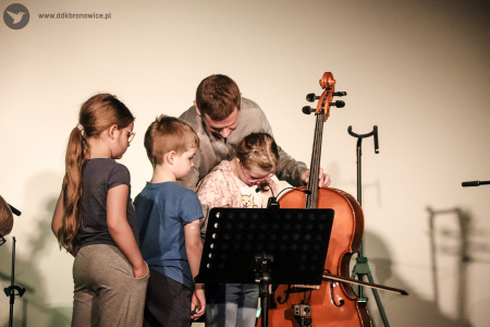 Kolorowe zdjęcie. Dzieci i mężczyzna na scenie. Mężczyzna uczy dziewczynkę grać na wiolonczeli. Inne dzieci się temu przyglądają.