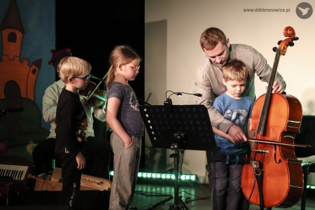 Kolorowe zdjęcie. Dzieci i mężczyzna na scenie. Mężczyzna uczy chłopca grać na wiolonczeli. Inne dzieci się temu przyglądają.