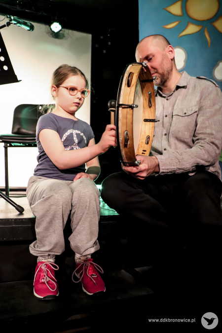 Kolorowe zdjęcie. Mężczyzna z dziewczynką siedzą na skraju sceny. Dziewczynka gra na bębenku obręczowym.