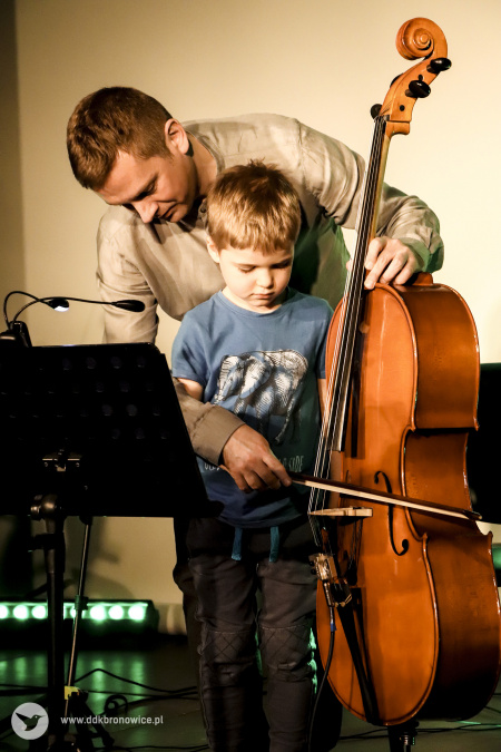 Kolorowe zdjęcie. Chłopiec i mężczyzna na scenie. Mężczyzna uczy chłopca grać na wiolonczeli.