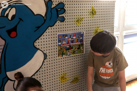 Kolorowe zdjęcie. Chłopiec przygląda się obrazkowi wiszącemu na ścianie. Po lewej stronie widoczna postać smerfa. Wokół obrazka wiszą żółte motyle.