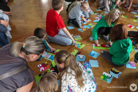 Kolorowe zdjęcie. Dzieci z rodzicami układają wspólnie wielkoformatowe puzzle na podłodze.