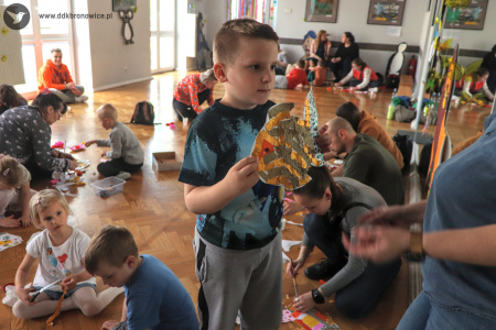 Kolorowe zdjęcie. Chłopiec trzyma przed animatorką w obu dłoniach wykonaną przez siebie rybkę z kartonu i złotej bibuły. W tle na podłodze siedzą rodzice z dziećmi i wykonują prace plastyczne.