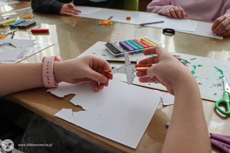 Dzieci robią z papieru i plasteliny ptaki. Dziewczynka na pierwszym planie zrobiła ptaka w kolorze białym z pomarańczowymi nogami i dziobem.