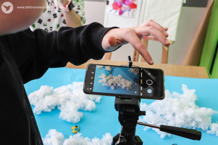 Na statywie telefon. Chłopiec robi zdjęcie dotykając ekranu. Fotografuje papierowe przedstawienie nieba z chmurami. Chmury zrobione z białej waty leżą na niebieskim papierze.