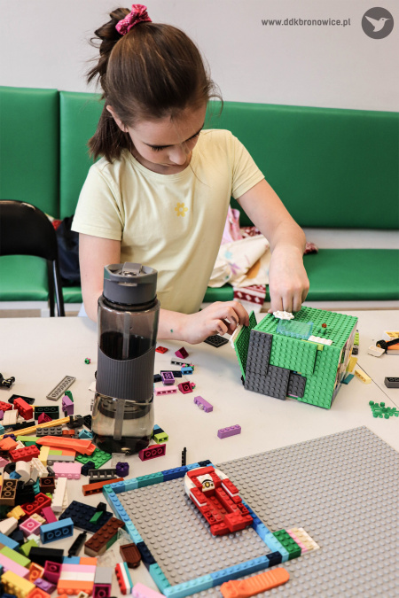 Dziewczynka bawi się klockami lego. Buduje zielony dom. Po lewej stronie stołu leżą wysypane klocki.