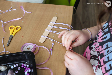 Na drewnianej podstawce, zawieszone fioletowe, opadające, sznureczki. Dziewczynka tworzy dalej swoją ozdobę.
