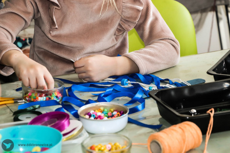 Dziewczynka tworzy swoją artystyczna ozdobę. Na stole leży niebieska wstążka, kolorowe małe kulki, nici.