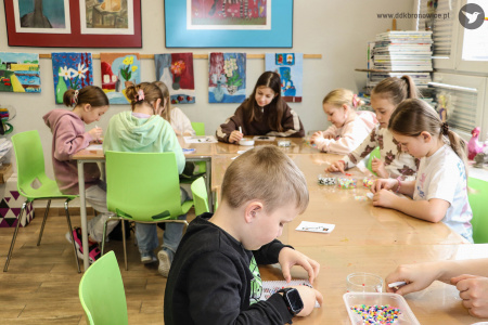 Grupa dzieci siedzi przy stole warsztatowym. Układają koraliki na plastikowych formach.