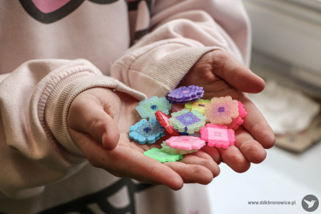 Dziewczynka pokazuje swoje kolorowe ozdoby, które trzyma w dłoniach. Przedstawiają kolorowe kwiatki.