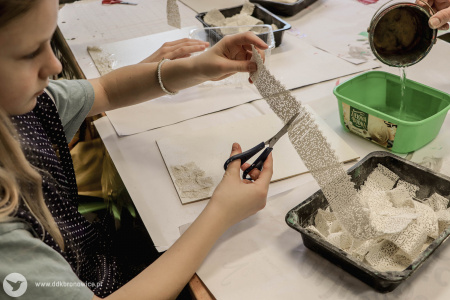 Dziewczynka wyciąga z czarnego pudełka podłużną siateczkę. Na białej kartce przed sobą ma przyklejony fragment takiej siatki.