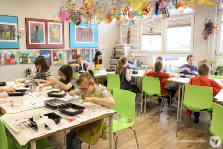 Grupa dzieci w skupieniu pracuje nad swoimi artystycznymi ozdobami. Siedzą na zielonych krzesłach.