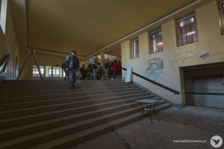 W budynku. Szeroki korytarz i schody. Odpada tynk i jest brudno. Grupa idzie  schodach. Ściany są pożółkłe.
