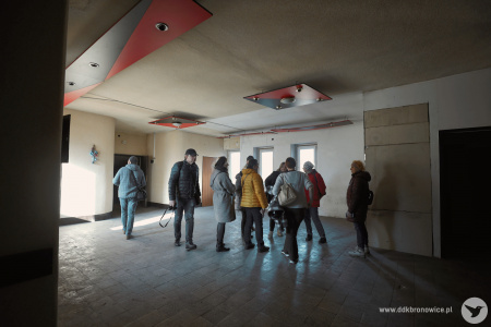 Grupa uczestników rozmawia w holu. Szara podłoga i ściany. Na suficie, czerwone lampy.