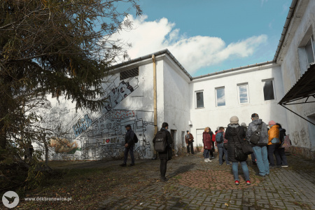 Biały piętrowy budynek. Na lewej ścianie grafiti. Grupa stoi przed budynkiem.