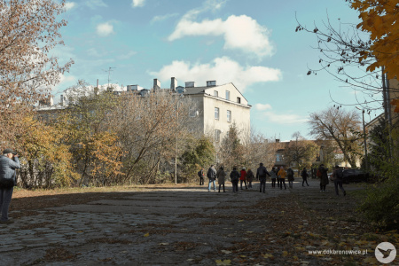 Stare budynki i drzewa. Zniszczony chodnik. Uczestnicy przemieszczają się. Kilka osób robi zdjęcia.