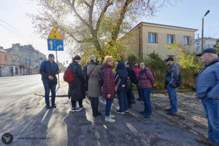 Na chodniku stoją uczestnicy. Obok ruchliwa ulica. Widać znak drogowy i drzewa za osobami.