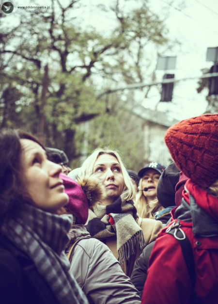 Spacer z przewodnikiem. Na pierwszym planie rozglądający się ludzie. Widać uczestniczkę, która z ciekawością patrzy w górę. W tle widać twarz przewodniczki. Ma założony mikrofon.