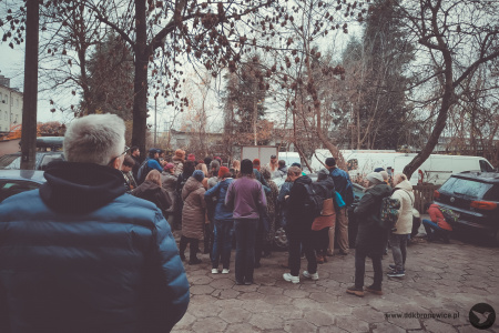 Pani przewodnik opowiada historię uczestnikom. Duża grupa ludzi stoi i słucha. Pochmurno. Na pierwszym planie mężczyzna w granatowej kurtce. W tle grupa słucha pani przewodnik.
