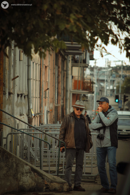Dwóch uczestników warsztatów fotograficznych rozmawia na chodniku. Obok kamienice. Na pierwszym planie, gałęzie drzewa.
