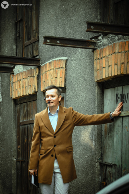 Przewodnik w brązowym płaszczu i błękitnej koszuli. Mówi do mikroportu. Opiera się jedną ręką o ścianę.