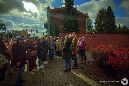 Spotkanie z przewodnikiem na placu. Obok ruchliwa ulica. Uczestnicy stoją po lewej stronie. Mają założone kurtki. Widać niebo i chmurę, która przysłoniła słońce.